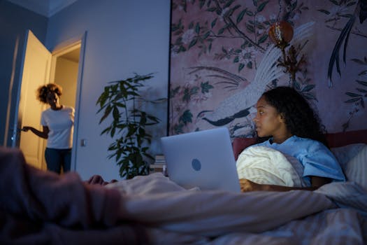 A teenager with a laptop in a dimly lit bedroom looks towards a parent in the doorway.