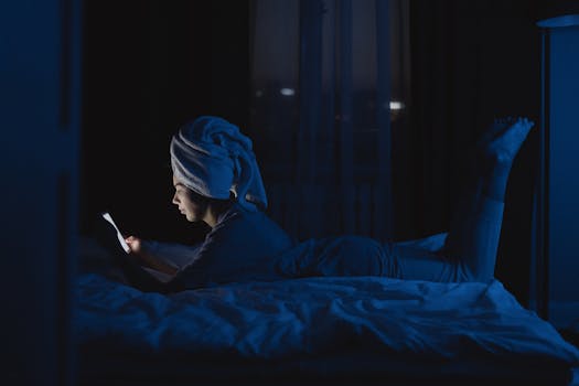 A woman lying in bed with a towel on her head, using a mobile phone at night.